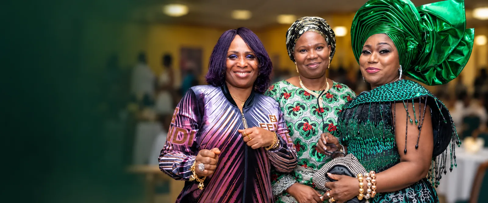 Three women in vibrant Yoruba attire smiling together at a community event.